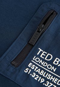Marineblåt stof med en lynlåslomme, der har et svart lynlåsgreb. Hvidt trykt tekst inkluderer "TED BAKER," "LONDON," og etableringsoplysninger.