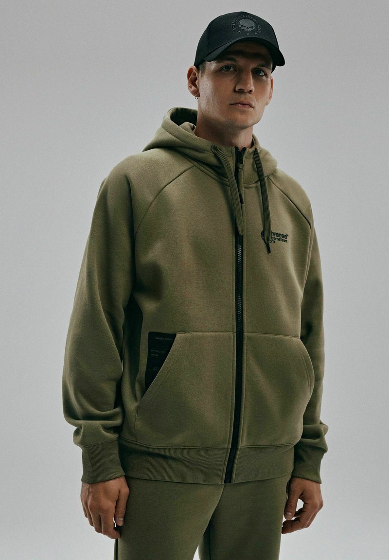 Oliwkowa bluza z kapturem zapinana na zamek, z przednią kieszenią, prążkowanymi mankietami i sznurkiem przy kapturze. Posiada czarną metkę na kieszeni. Gładka tekstura.