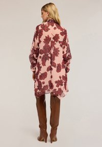 Chiffonbluse mit pinkem Grund und großem roten Blumenmuster; hat lange Ärmel und einen asymmetrischen Saum. Trägt man mit braunen Overkneestiefeln.