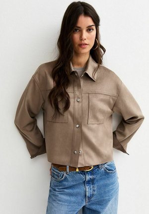 Kobieta o długich ciemnych włosach, ubrana w beżową kurtkę zapinaną na guziki, złote kolczyki koła oraz niebieskie dżinsy, stoi oparta o białą ścianę z rękami za plecami.