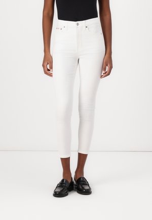Person trägt weiße, gekürzte Skinny-Jeans, schwarzen ärmellosen Top und schwarze glänzende Loafers und steht vor einem einfarbigen weißen Hintergrund.