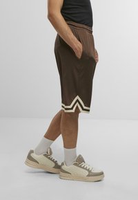 Shorts de sport marron avec une surface texturée, dotés de rayures blanches en chevron près de l'ourlet. Associés à des baskets de couleur claire et des chaussettes blanches.