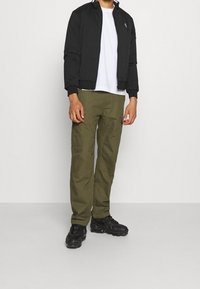 Veste bombardier noire, t-shirt blanc, pantalon cargo vert olive et baskets noires. Caractéristiques incluant des poignets côtelés et plusieurs poches sur le pantalon.