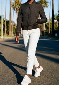 Personne marchant dans une rue ensoleillée portant une veste noire, un pantalon blanc et des baskets blanches avec des palmiers visibles en arrière-plan.