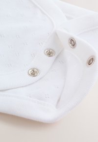 Tissu blanc pour vêtement bébé avec de petits points texturés, comprenant deux paires de boutons-pression argentés sur les bords côtelette.