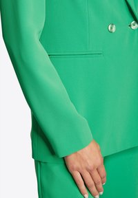 Zielony, dopasowany garnitur o gładkiej fakturze. Marynarka posiada podwójne guziki i fason slim z boczną kieszenią, podkreślając nowoczesny wygląd.