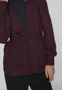Femme portant un cardigan tricoté violet foncé noué à la taille sur une tenue noire, montrée des épaules aux hanches.