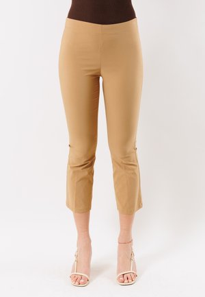 Donna che indossa pantaloni beige corti e una maglia marrone, abbinati a sandali bianchi con tacco alto e punta aperta, e una cavigliera alla caviglia sinistra.