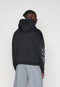 Μαύρη κουκούλα φούτερ με cropped σχέδιο, υφή υφάσματος, oversized εφαρμογή και λευκές γραφικές λεπτομέρειες στους μανίκους.