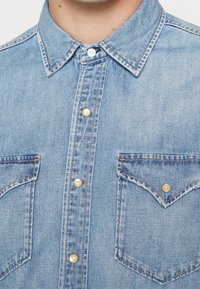 Ljusblå denimskjorta med knappförsedd framsidan, spetsig krage, två bröstfickor och pärlkapslade knappar. Har en klassisk westerndesign.