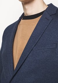 Blazer blu navy realizzato in tessuto texture con rever a punta e taschino sul petto, indossato sopra un maglione marrone con scollatura nera.
