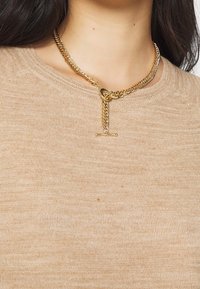 Guldhalsband med en tvistad design, med en lås med spänne och en avlång medaljong, bärs över en ljus beige texturerad tröja.