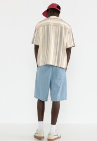 Chemise à manches courtes rayée beige et grise, associée à un short en denim bleu clair. Les accessoires comprennent une casquette rouge et des baskets beiges.