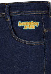 Jeans en denim bleu foncé avec des coutures jaunes, dotés d'une poche et d'un logo brodé "homeboy x-tra" en orange et bleu sur le devant.