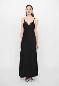 Musta, nilkkapituisen helman mekko ohuillaolkaimilla, syvällä V-aukolla, epäsymmeetrisellä muotoilulla, sileällä tekstuurilla ja elegantilla laskoksella.