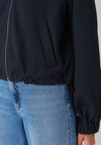 Veste zippée marine avec une texture côtelée, des poignets élastiques et une poche latérale. Portée avec un jean bleu clair pour un look décontracté.