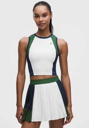 Atletisk kvinde iført en ærmeløs, farveblokkeret crop top og plisseret nederdel i hvid, grøn og marineblå, stående mod en ensfarvet baggrund.