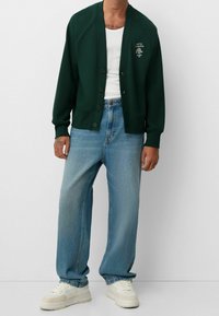 Sudadera verde oscura con cierre de botones y puños acanalados, combinada con jeans de pierna recta azul claro y zapatillas blancas.