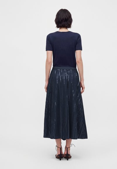Femme aux cheveux courts et foncés portant un haut bleu marine à manches courtes et une jupe midi plissée foncée avec des rayures verticales brillantes, debout dos à la caméra.