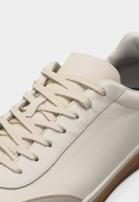 Primo piano di una sneaker in pelle color crema con lacci testurizzati e suola in gomma marrone chiaro su sfondo bianco.