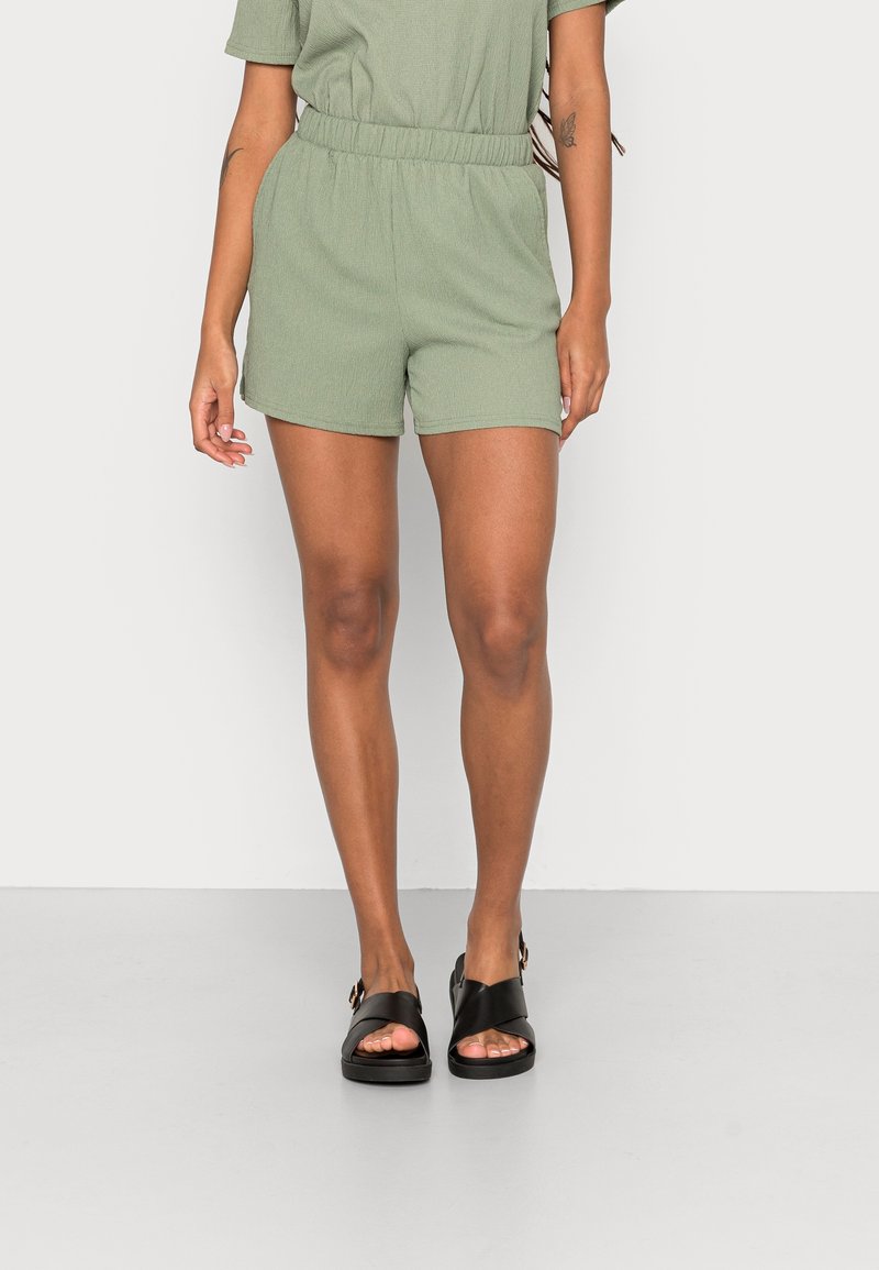 Combinaison courte verte texturée avec taille élastique, associée à des sandales noires à brides croisées. Accent sur le confort et le style décontracté.