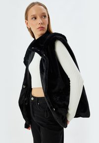 Schwarze Kunstpelzweste mit lässiger Passform, Druckknopfverschluss und seitlichen Taschen, kombiniert mit einem weißen gerippten Cropped-Top und schwarzen Jeans.