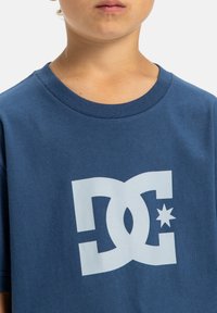 Blaues Baumwoll-T-Shirt mit Rundhalsausschnitt, das ein großes weißes DC-Logo und einen Sternakzent auf der Vorderseite zeigt. Weicher Stoff, kurze Ärmel.