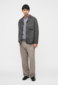 Γκρι σακάκι από μαλλί με ένα τσέπη στο στήθος, κούμπωμα με κουμπιά και γιακά με σκίσιμο, σε συνδυασμό με ανοιχτό μωβ πουλόβερ και μπεζ παντελόνια.