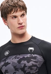 Czarna sportowa koszulka o dopasowanym kroju, z szarym wzorem kamuflażu na torsie oraz białymi logo na ramionach.