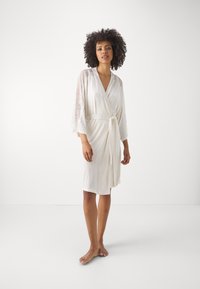 Robe avvolgente bianco con maniche in pizzo a tre quarti, dotato di cintura da annodare e texture liscia. Indossato a piedi nudi su uno sfondo neutro.