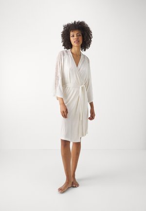 Robe avvolgente bianco con maniche in pizzo a tre quarti, dotato di cintura da annodare e texture liscia. Indossato a piedi nudi su uno sfondo neutro.