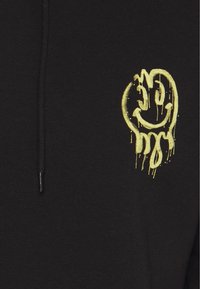 Svart sweatshirt med ett stort gult leende ansikte med droppar, tillverkat av mjukt tyg, standard passform och huva med dragsko.