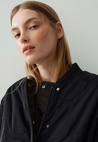Veste bomber noire matelassée avec col côtelé, boutons-pression et fermeture éclair, mise en valeur sur un modèle devant un arrière-plan clair.