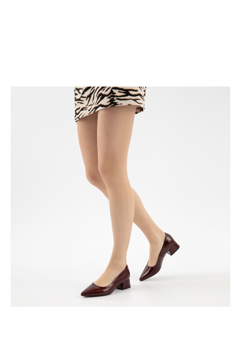 Donna che indossa calze velate color carne e scarpe bordeaux con tacco basso e punta appuntita, in piedi con una gamba incrociata davanti all'altra.