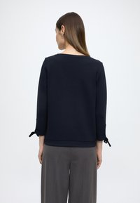 Granatowa prążkowana bluza z okrągłym dekoltem i rękawami do połowy przedramienia z wiązanymi mankietami, w połączeniu z szarymi luźnymi spodniami.