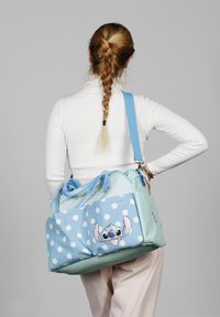 Bolsa de lunares azules con un gráfico del personaje Stitch, que cuenta con dos bolsillos laterales, un diseño de chevrón en la parte superior y correas ajustables azules.