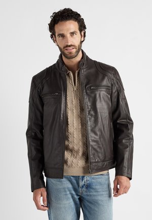 Brązowa skórzana kurtka z kołnierzem, z dwoma zamykanymi na zamek kieszeniami na piersi oraz subtelnymi detalami stębnowania. Noszona na beżowym swetrze robionym na drutach.