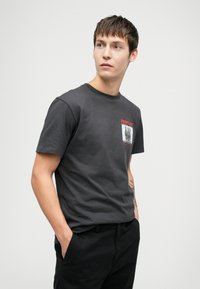 Νεαρός άνδρας με σκούρα κοντά μαλλιά, φορώντας μαύρο μπλουζάκι με γραφικά και μαύρο παντελόνι, με τα χέρια στις τσέπες και κοιτάζοντας λίγο στο πλάι.