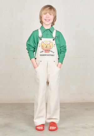 Groene lange mouwen shirt onder denim tuinbroek met een hondenafbeelding. Crème-kleurige stof en een relaxte pasvorm. Rode sandalen met branding.