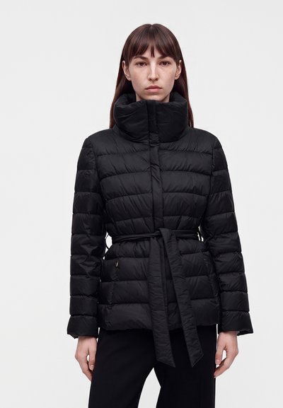 Femme portant une veste matelassée noire avec un col montant et une ceinture nouée, debout devant un fond clair uni.