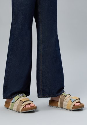 Piedi con sandali aperti beige, verdi e gialli con cinturini regolabili abbinati a jeans a gamba larga blu scuro su uno sfondo neutro.