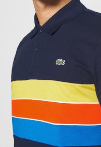 Námořnicky modré polo tričko s horizontálními pruhy v žluté, oranžové, modré a zelené barvě, s malým vyšívaným logem na hrudi.