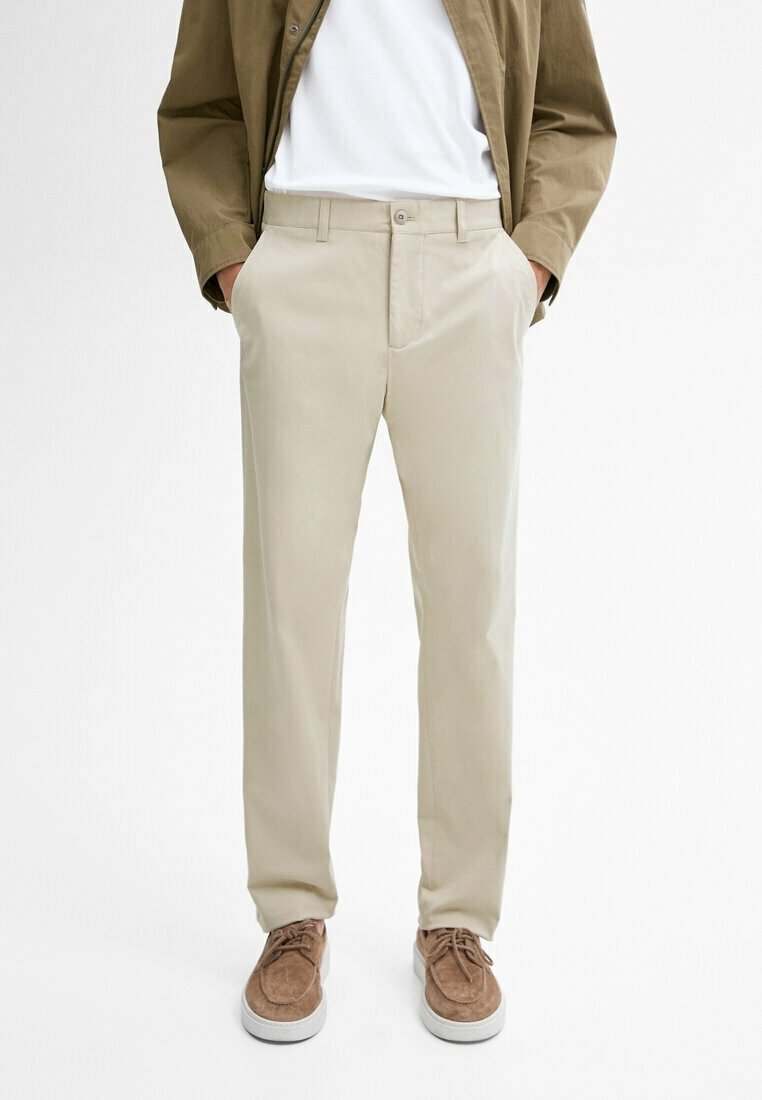 Pantalon beige à jambes droites porté avec des chaussures en daim marron, un t-shirt blanc et une veste marron clair, mains dans les poches, debout sur fond blanc.