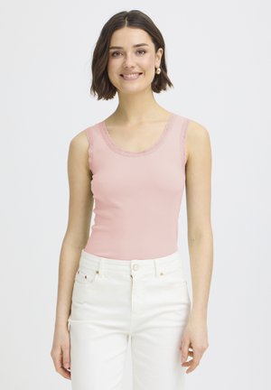 Noor naine lühikeste tumejuustega, kes naeratab, kannab roosat varrukateta topi pitsist äärisega ja valgeid kõrge vöökohaga pükse.