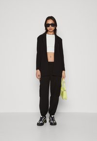 Fekete, oversized blézer, fehér cropped felső, fekete cargo nadrág, fekete és sárga sportcipők, valamint egy kis világos zöld kézitáska. Napszemüveg viselve.