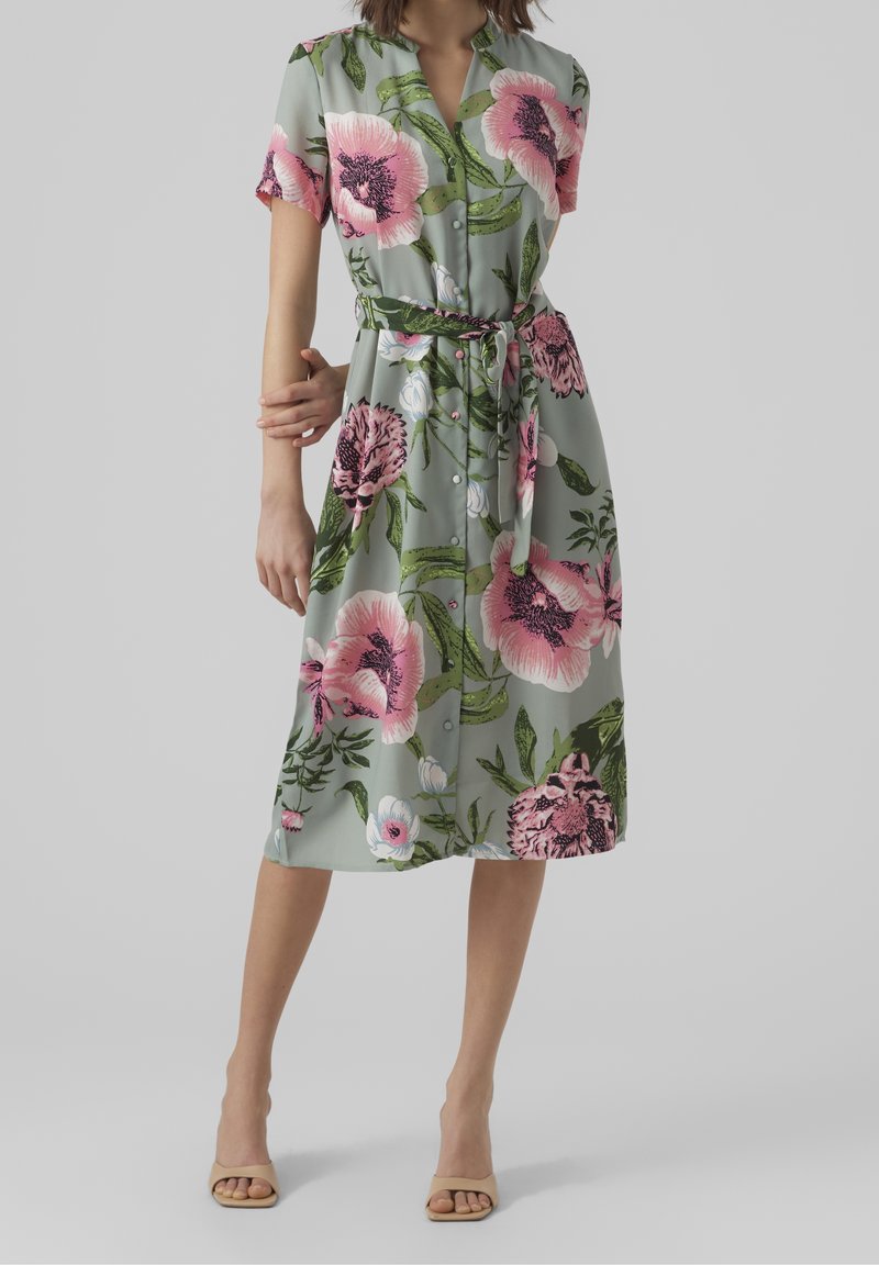 Donna che indossa un vestito midi con cintura e bottoni, con maniche corte, realizzato in un tessuto verde con grandi fiori rosa e bianchi.