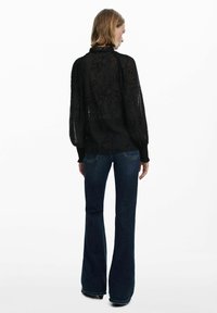 Blusa nera trasparente con ricami floreali, colletto alto e maniche lunghe a sbuffo. Abbinata a jeans a zampa di elefante blu scuro.