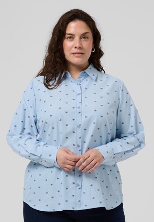 Woman with dark curly hair wears a light blue button-up shirt patterned with small black bows, standing against a gray background.