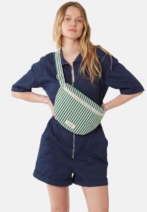 Femme aux longs cheveux blonds portant une combinaison bleu marine zippée et un sac bandoulière rayé vert et blanc.