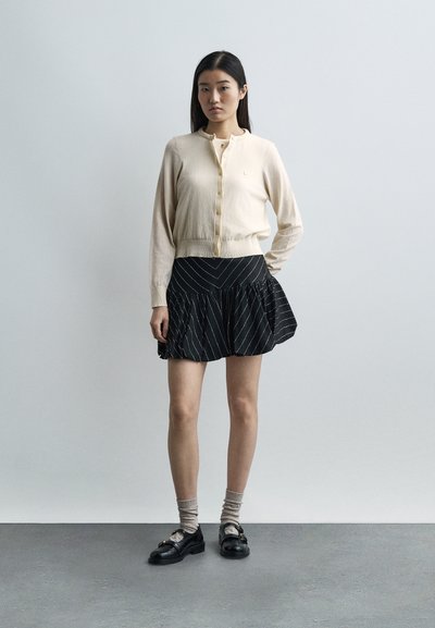 Γυναίκα που φοράει μπεζ ζακέτα, μαύρη φούστα με λευκές ρίγες, μπεζ κάλτσες και μαύρα παπούτσια, στέκεται μπροστά σε ένα απλό ανοιχτό γκρι φόντο.